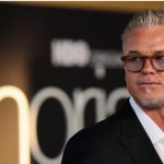 A professional headshot of actor Eric Dane at a red carpet event. He is wearing a black suit jacket over an unbuttoned white shirt and amber-tinted glasses. The background is blurred with a glowing "Euphoria" sign visible. Overlaying the image is a fake news headline claiming he passed away at age 53 after a battle with ALS.
