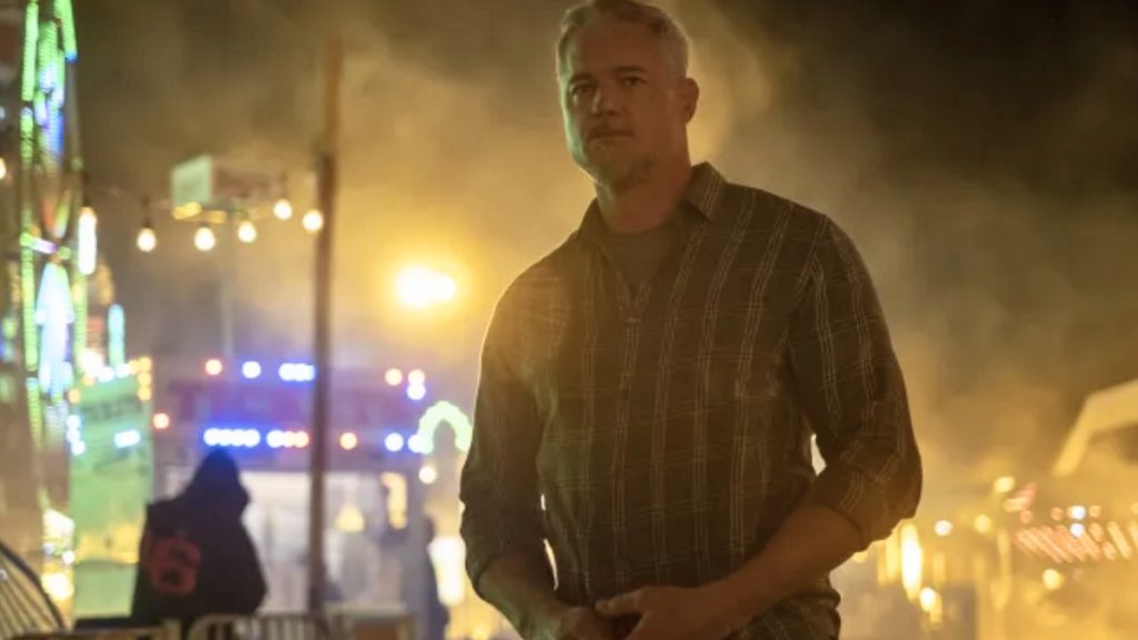 A cinematic shot of Eric Dane as his character Cal Jacobs from the show Euphoria. He is standing outdoors at night at a carnival, wearing a plaid button-down shirt. The background is filled with warm, glowing lights and hazy smoke from the fairground. A fake caption at the top suggests this is a scene from "Season 3."