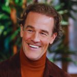 A medium close-up shot of actor James Van Der Beek, known for his role in "Dawson's Creek." He is smiling warmly, looking slightly off-camera to the right. He has wavy light brown hair and a light stubble. He is wearing a brown suede jacket over a rust-colored turtleneck sweater. The background is softly blurred, showing hints of green indoor plants and warm lighting.