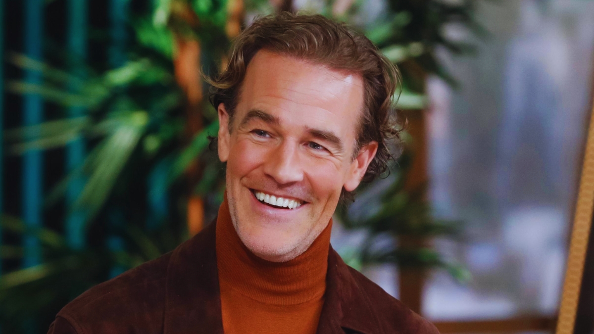 A medium close-up shot of actor James Van Der Beek, known for his role in "Dawson's Creek." He is smiling warmly, looking slightly off-camera to the right. He has wavy light brown hair and a light stubble. He is wearing a brown suede jacket over a rust-colored turtleneck sweater. The background is softly blurred, showing hints of green indoor plants and warm lighting.