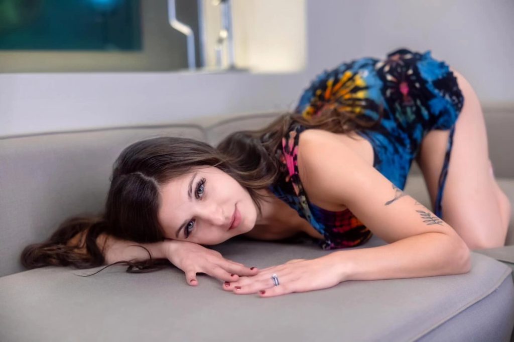 Leah Gotti reclining on a light grey sofa, leaning forward on her elbows with her head resting near her hands. She is wearing a colorful tie-dye patterned dress in blue, orange, and black. She has a soft smile and is looking toward the lens, with her long dark hair cascading over her shoulder and a visible tattoo on her right forearm.