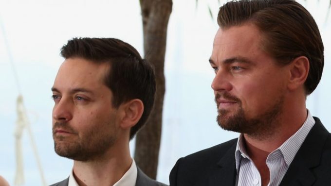 Actors Leonardo DiCaprio and Tobey Maguire looking toward the side during a Super Bowl party; DiCaprio is sporting a beard and Maguire is in a white dress shirt.