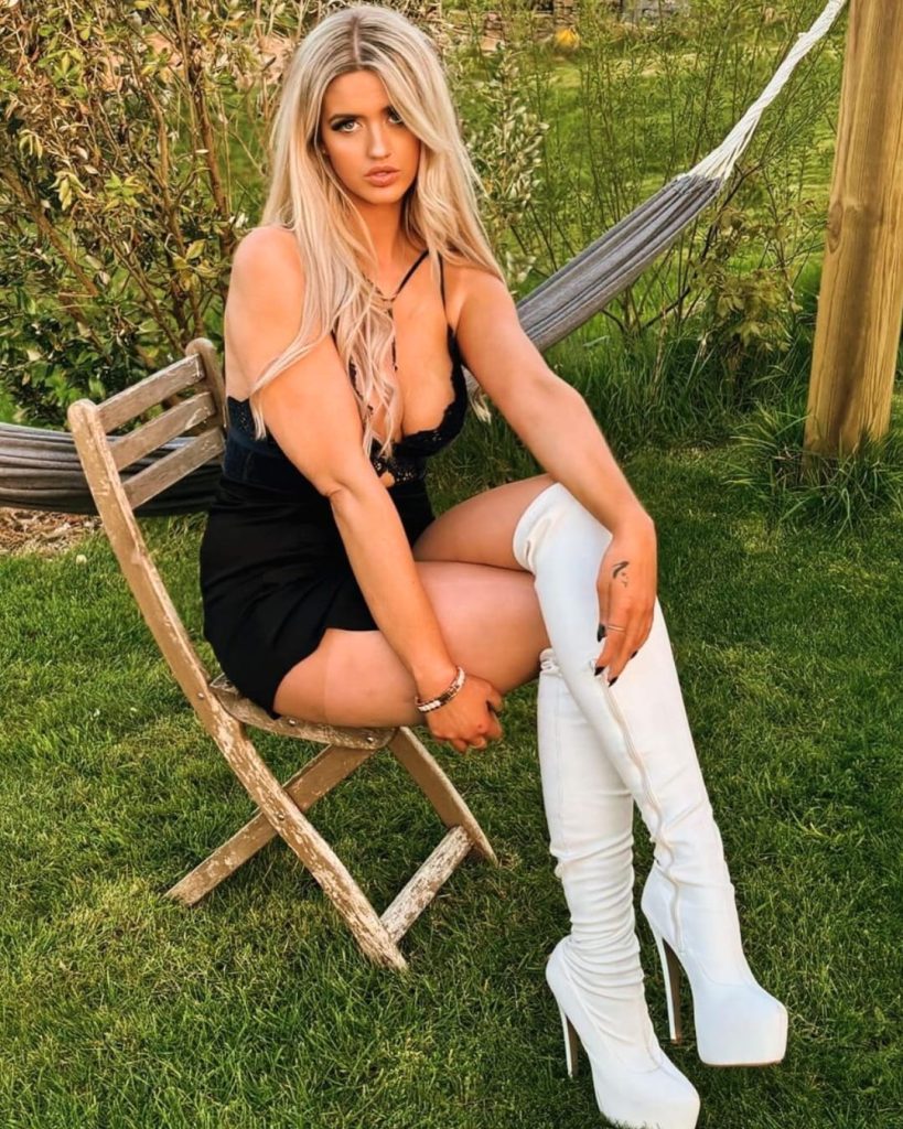 Tall model Lizzy Jade Groombridge sitting on a wooden chair outdoors, wearing a black lace bodysuit, mini skirt, and white platform thigh-high boots.