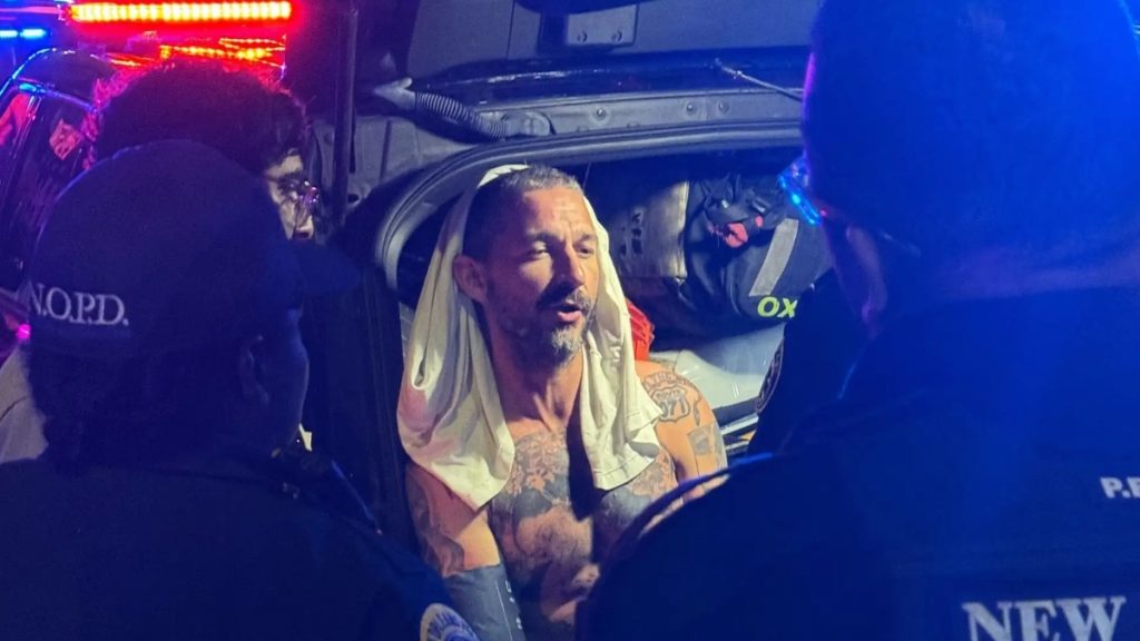 A candid, low-light photograph showing Shia LaBeouf shirtless and heavily tattooed, seated in the back of a vehicle surrounded by police officers. He has a white t-shirt draped over his head and appears to be speaking or shouting. The scene is illuminated by the harsh blue and red flashing lights of emergency vehicles. The back of a New Orleans Police Department (N.O.P.D.) officer's hat is visible in the foreground.