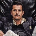 A dramatic, high-contrast portrait of the drug lord known as "El Mencho." He is seated in a large, ornate black leather chair, wearing a dark pinstriped suit over a light-colored button-down shirt. He has a serious expression, a thin mustache, and is leaning forward with his arms resting on the chair's armrests. The image is watermarked with the text "VICESNOB" in the upper right corner.