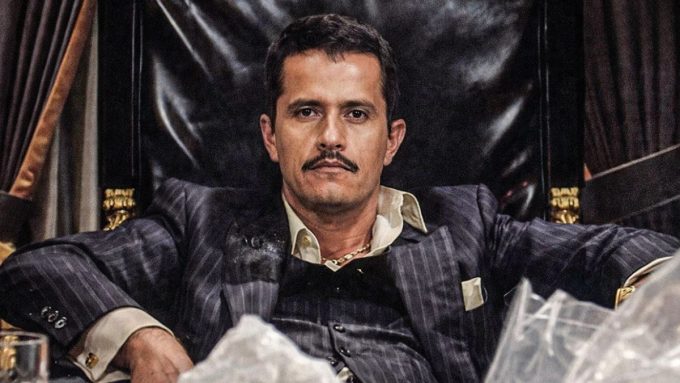 A dramatic, high-contrast portrait of the drug lord known as "El Mencho." He is seated in a large, ornate black leather chair, wearing a dark pinstriped suit over a light-colored button-down shirt. He has a serious expression, a thin mustache, and is leaning forward with his arms resting on the chair's armrests. The image is watermarked with the text "VICESNOB" in the upper right corner.