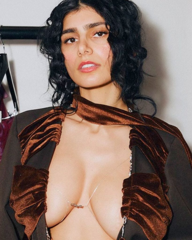 Mia Khalifa wearing a brown velvet-trimmed jacket with a delicate body chain accessory.