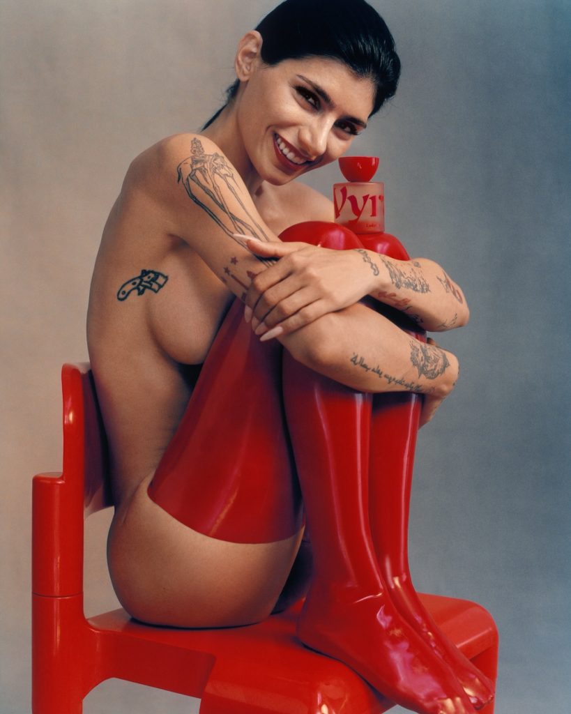 A high-fashion studio shot of Mia Khalifa wearing red latex boots, posing artistically with a perfume bottle, following a viral Mia Khalifa bikini shoot.