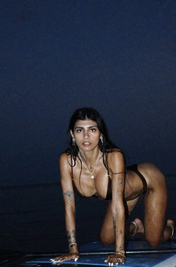 Mia Khalifa posing on all fours on a paddleboard at night, wearing a dark-colored Mia Khalifa bikini and layered gold jewelry.