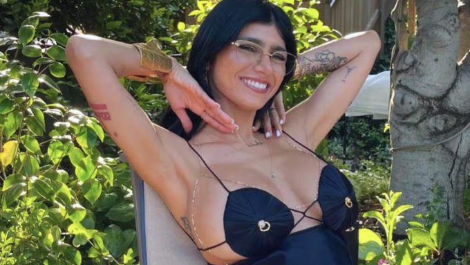 A smiling Mia Khalifa with dark hair and glasses poses with her arms behind her head in a sunny garden setting, wearing a black decorative top and gold jewelry. From the Mia Khalifa Birthday 2026 carousel.