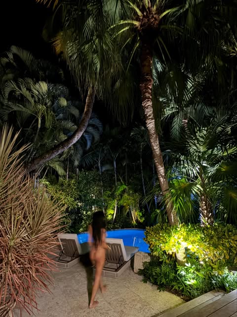 A blurred, artistic long-exposure shot of a person walking toward a glowing blue swimming pool at night, surrounded by lush tropical palm trees. Part of a Mia Khalifa Birthday social media collection.