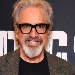 A more recent headshot of actor Robert Carradine smiling in front of a dark backdrop with white lettering. He is sporting grey hair, a matching grey goatee, and thick-rimmed black glasses, while wearing a dark t-shirt under a grey herringbone blazer.