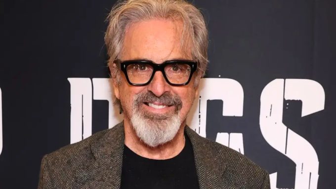 A more recent headshot of actor Robert Carradine smiling in front of a dark backdrop with white lettering. He is sporting grey hair, a matching grey goatee, and thick-rimmed black glasses, while wearing a dark t-shirt under a grey herringbone blazer.