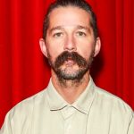 A medium close-up, front-facing portrait of actor Shia LaBeouf against a vibrant red curtain backdrop. He has a serious, neutral expression, short dark hair slicked back, and a very thick, prominent handlebar mustache paired with a light beard. He is wearing a simple, tan or light khaki button-down shirt.