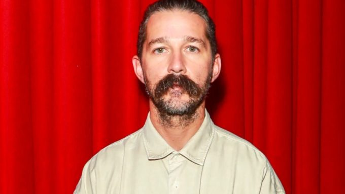 A medium close-up, front-facing portrait of actor Shia LaBeouf against a vibrant red curtain backdrop. He has a serious, neutral expression, short dark hair slicked back, and a very thick, prominent handlebar mustache paired with a light beard. He is wearing a simple, tan or light khaki button-down shirt.