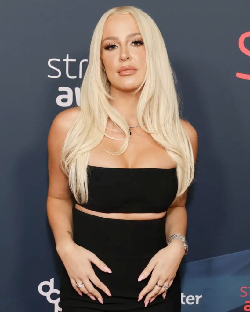 Tana Mongeau wearing a black strapless crop top and high-waisted skirt with long platinum blonde hair.