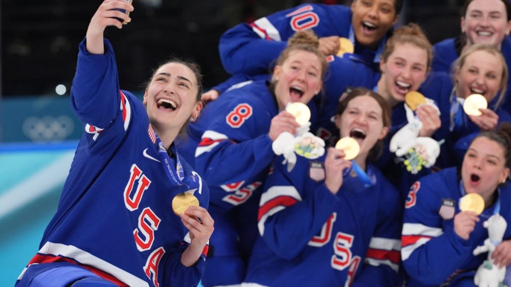 A close-up of US Women's Hockey Team players celebrating their 2-1 overtime victory against Canada at the Milano Cortina 2026 Games, paired with a headline regarding their declined White House invitation.