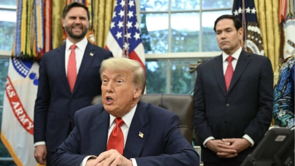 A medium shot of Donald Trump seated at the Resolute Desk in the Oval Office, speaking with an expressive gesture. Behind him stand JD Vance on the left, smiling, and Marco Rubio on the right, maintaining a serious expression. All three men are wearing dark suits and red ties. An American flag and a U.S. Army flag are visible in the background.