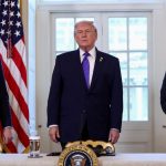 A wide shot of JD Vance, Donald Trump, and Marco Rubio standing side-by-side behind a long table featuring the Presidential Seal. Donald Trump stands in the center wearing a purple tie, while JD Vance (left) and Marco Rubio (right) stand at their respective stations, indicated by nameplates. Vance is looking toward Trump with a slight smile, while Rubio looks forward with a neutral expression.