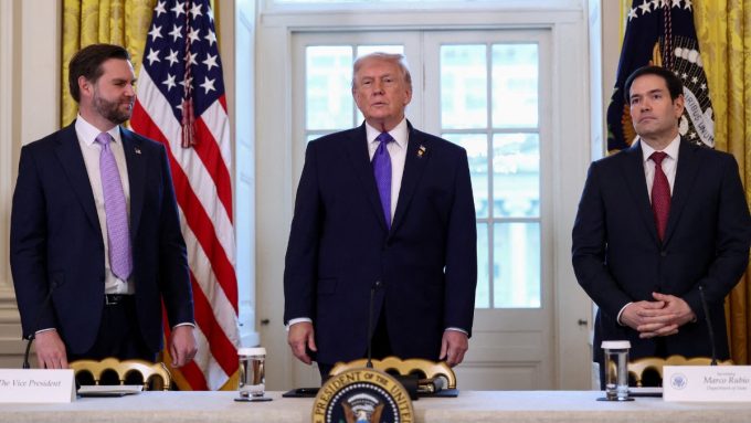 A wide shot of JD Vance, Donald Trump, and Marco Rubio standing side-by-side behind a long table featuring the Presidential Seal. Donald Trump stands in the center wearing a purple tie, while JD Vance (left) and Marco Rubio (right) stand at their respective stations, indicated by nameplates. Vance is looking toward Trump with a slight smile, while Rubio looks forward with a neutral expression.