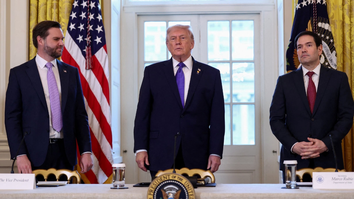 A wide shot of JD Vance, Donald Trump, and Marco Rubio standing side-by-side behind a long table featuring the Presidential Seal. Donald Trump stands in the center wearing a purple tie, while JD Vance (left) and Marco Rubio (right) stand at their respective stations, indicated by nameplates. Vance is looking toward Trump with a slight smile, while Rubio looks forward with a neutral expression.