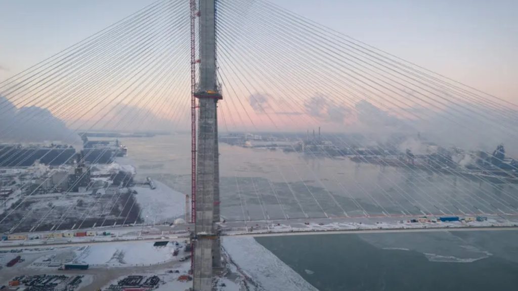 A wide, atmospheric aerial shot of the Gordie Howe International Bridge during winter. The scene shows the bridge’s tall concrete pylons and radiating stay cables stretching across a partially frozen river. The surrounding industrial landscape is covered in snow, with plumes of white steam rising from nearby factory stacks against a pale, hazy sky at dusk or dawn.