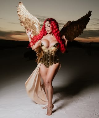 Veronica Melendez Coronado with long, curly red hair dressed in a metallic gold corset and flowing skirt. She is wearing large gold and black wings, posing in a desert setting at dusk.
