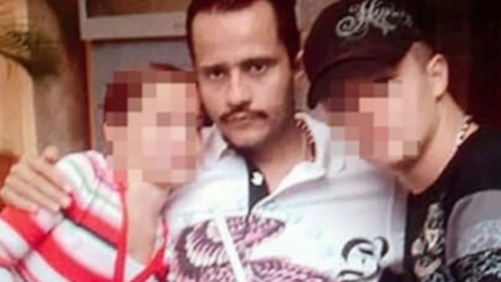 A grainy, candid photograph showing three people. In the center is a man with a mustache, identified as a younger El Mencho. He has his arms wrapped around two other individuals—a person on the left in a striped shirt and a person on the right wearing a black baseball cap.