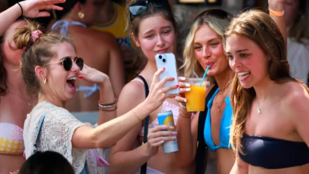 A group of young women celebrating at a lively spring break 2026 event. One woman in sunglasses laughs while holding a phone to take a selfie, surrounded by others in swimwear enjoying drinks and the festive atmosphere.