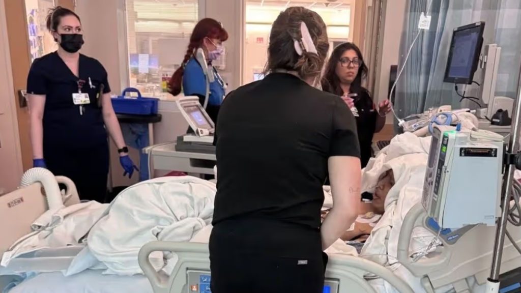 A wide shot of a hospital room where Alex Duong is being treated by several medical staff members. Nurses in dark scrubs and masks are seen monitoring equipment and tending to Alex, who is lying in the hospital bed surrounded by medical monitors and IV poles.