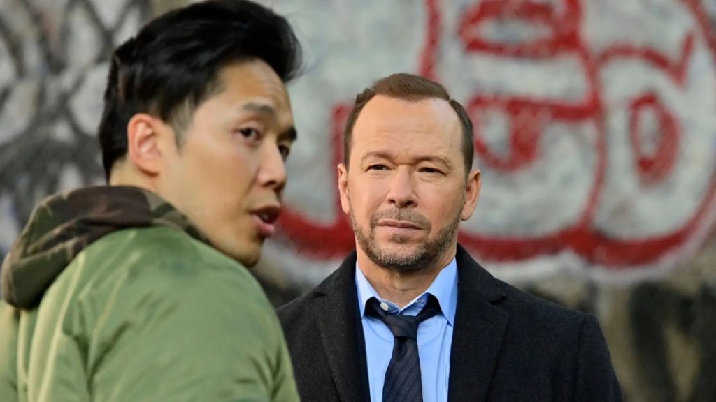 A television still from the series "Blue Bloods" featuring Alex Duong in his recurring role as Sonny Le. He is seen in a green jacket in the foreground, speaking to Donnie Wahlberg’s character, Danny Reagan, who is listening intently in a dark coat and tie.