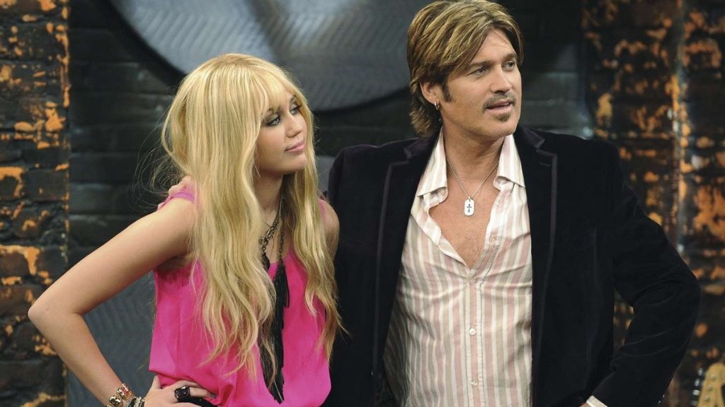 A classic scene featuring Miley Cyrus as Hannah Montana and Billy Ray Cyrus as Robby Ray Stewart, shared in celebration of the Hannah Montana 20th Anniversary; Miley wears her signature blonde wig and a bright pink top while Billy Ray stands beside her in a striped shirt and black blazer.