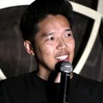 Gemini said This is a close-up photograph of comedian Alex Duong, taken during a performance at a comedy club. He is smiling and looking to his left, with the mic in his right hand. Alex, who has short black hair, is wearing a black crew-neck t-shirt. He has a subtle mustache and his teeth are visible as he grins.