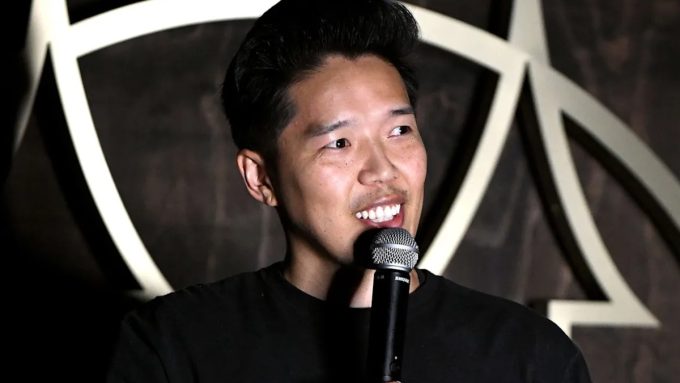 Gemini said This is a close-up photograph of comedian Alex Duong, taken during a performance at a comedy club. He is smiling and looking to his left, with the mic in his right hand. Alex, who has short black hair, is wearing a black crew-neck t-shirt. He has a subtle mustache and his teeth are visible as he grins.