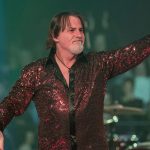 Tommy DeCarlo performing in a vibrant, red sequined button-down shirt. He is smiling and pointing toward the audience with one hand while holding a microphone in the other. The stage lighting features green and blue hues, highlighting his role as an iconic voice in rock.