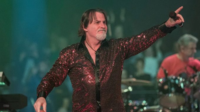 Tommy DeCarlo performing in a vibrant, red sequined button-down shirt. He is smiling and pointing toward the audience with one hand while holding a microphone in the other. The stage lighting features green and blue hues, highlighting his role as an iconic voice in rock.