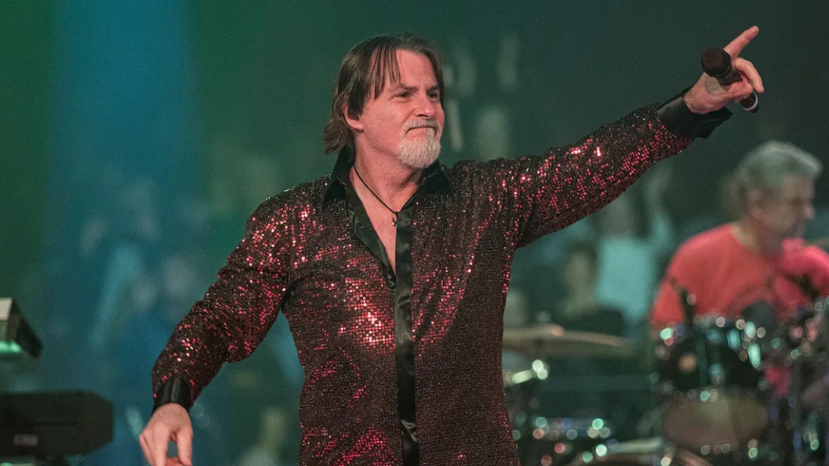 Tommy DeCarlo performing in a vibrant, red sequined button-down shirt. He is smiling and pointing toward the audience with one hand while holding a microphone in the other. The stage lighting features green and blue hues, highlighting his role as an iconic voice in rock.