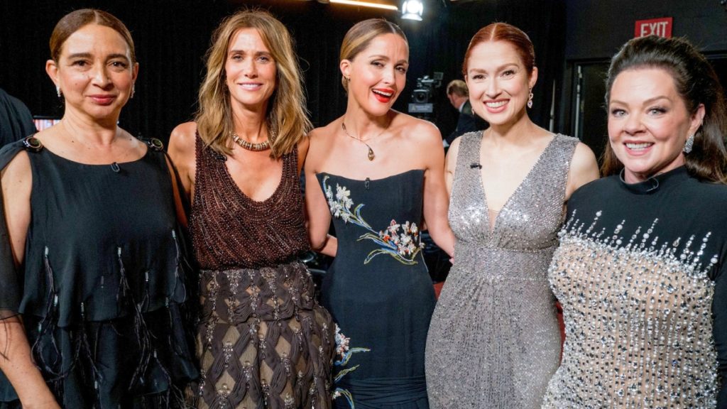 A group photo featuring the cast of Bridesmaids reunited backstage at the 2026 Oscars. From left to right: Maya Rudolph in a black fringed top, Kristen Wiig in a beaded brown gown, Rose Byrne in a strapless black dress with floral embroidery, Ellie Kemper in a shimmering silver V-neck dress, and Melissa McCarthy in a black gown with silver sequined detailing. They are all smiling and posed closely together.