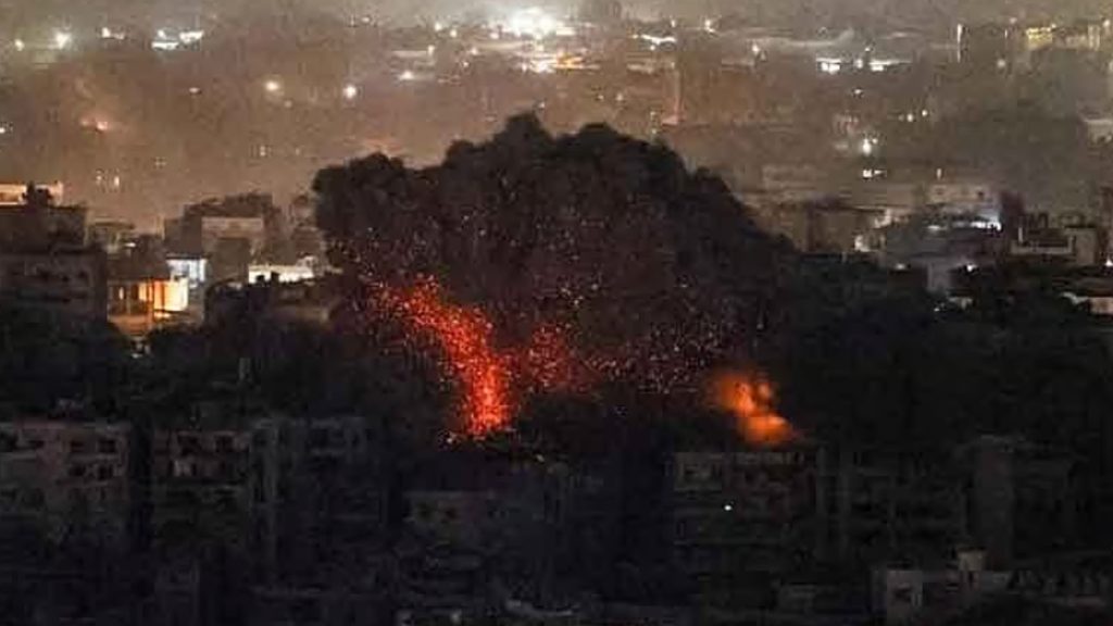 A grainy nighttime photograph showing a large, fiery explosion and a plume of dark smoke rising above a densely populated city during the Israel Iran War. Small fires are visible on the rooftops of nearby residential buildings