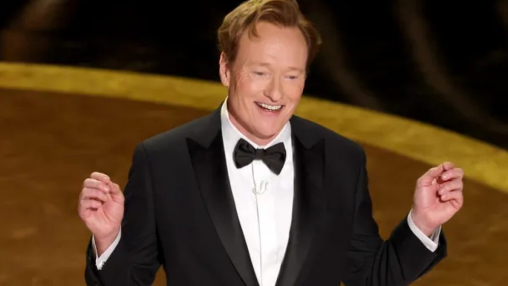 Conan O'Brien smiling on stage while hosting the Oscars 2026. He is wearing a classic black tuxedo with a black bow tie and a white dress shirt, captured in an upbeat moment during the ceremony broadcast.