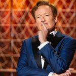 A medium shot of Conan O'Brien hosting the 2026 Oscars. He is wearing a sharp navy blue tuxedo with black velvet lapels and a black bowtie. He has a thoughtful expression with one finger held to his lips, posing against a geometric, golden-lit background.