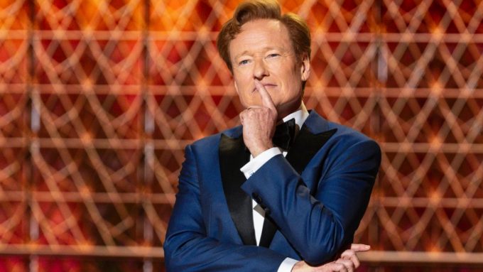 A medium shot of Conan O'Brien hosting the 2026 Oscars. He is wearing a sharp navy blue tuxedo with black velvet lapels and a black bowtie. He has a thoughtful expression with one finger held to his lips, posing against a geometric, golden-lit background.