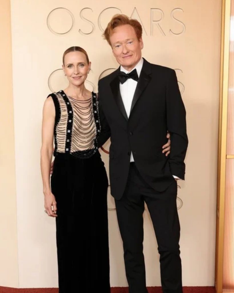 A photograph of Conan O'Brien and his wife, Liza Powel, posing together on a red carpet at an Oscars event. They are standing close, smiling at the camera. O'Brien, tall with his signature red hair, is wearing a black tuxedo with a black bow tie. Powel is wearing a long black sleeveless dress with a distinctive semi-sheer bodice featuring cascading rows of dark beads and silver embellishments. Her hair is pulled back in a neat, low bun. They stand against a light beige wall with the embossed "OSCARS" logo and a vertical gold-slatted partition. The red carpet is visible at their feet.