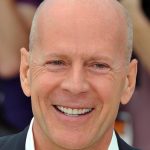 A close-up headshot of actor Bruce Willis smiling during a public appearance. The image is often used to accompany health updates or to address recurring rumors regarding his well-being.