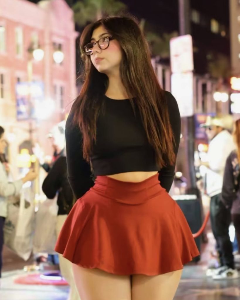 Young woman in a black top and red skirt stands confidently on a bustling city street at night, illuminated by colorful lights and surrounded by pedestrians.