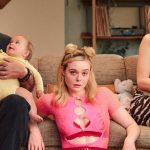 Elle Fanning sits on a couch wearing a pink mesh top, flanked by a bearded man holding a baby and a woman in a leopard-print dress, in a scene from Margo's Got Money Troubles.