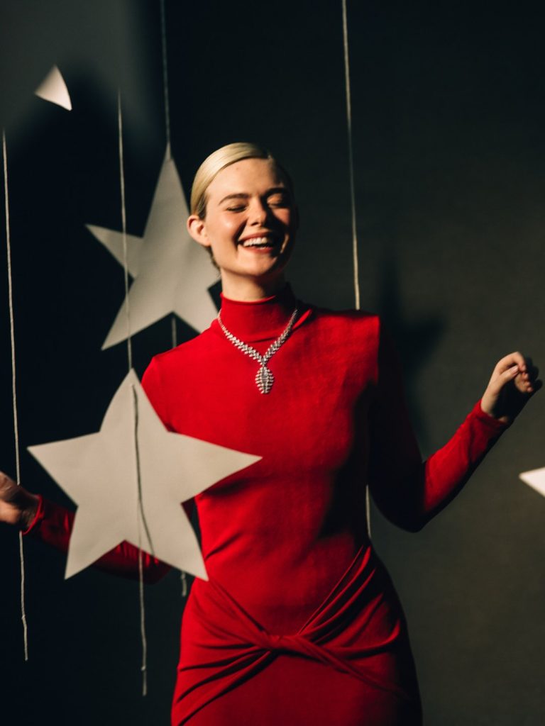 Elle Fanning posing for Margo's Got Money Troubles, wearing a red gown and luxury jewelry amidst a starry backdrop.