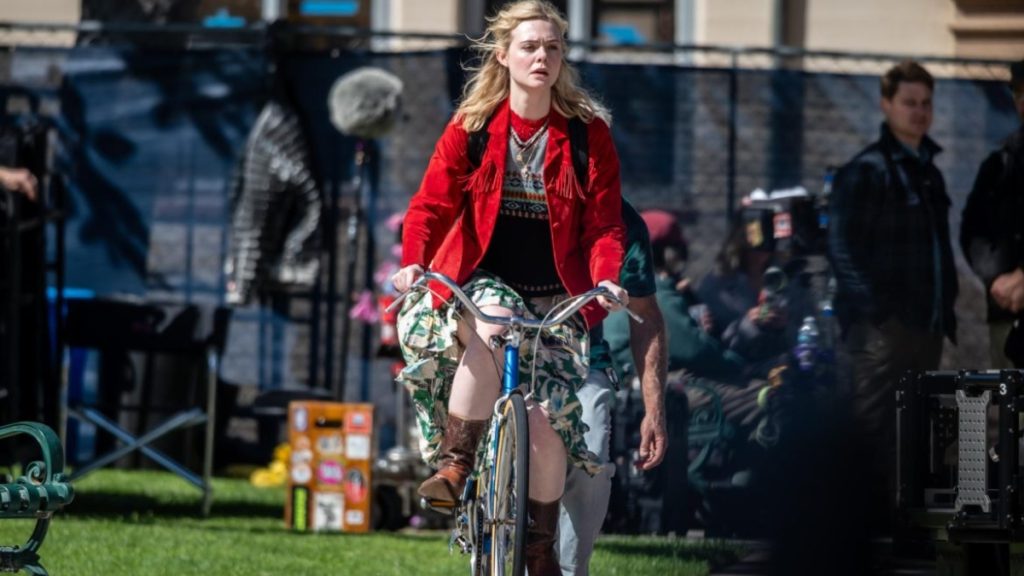 Behind-the-scenes look at Elle Fanning filming Margo's Got Money Troubles on location, featuring her character riding a bike in a red jacket.