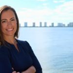 Emily Gregory smiling confidently in a navy blue dress, posed against a scenic waterfront backdrop with a city skyline on the distant horizon.