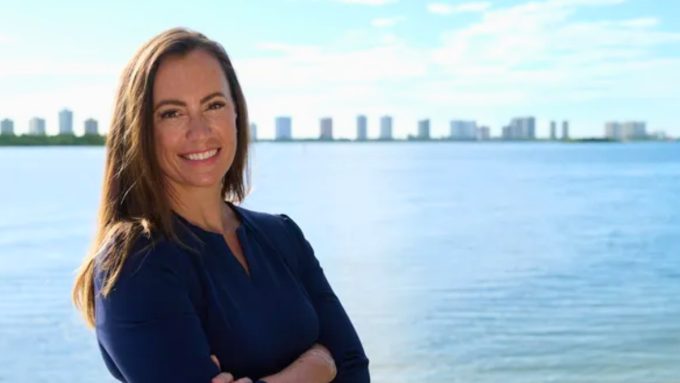 Emily Gregory smiling confidently in a navy blue dress, posed against a scenic waterfront backdrop with a city skyline on the distant horizon.